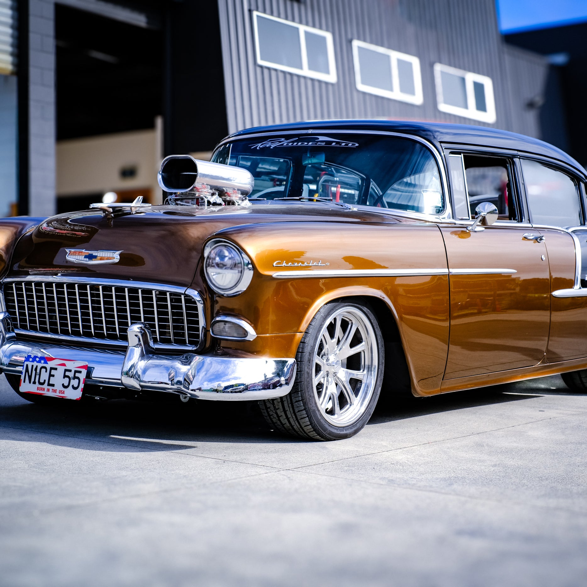 A customized 1957 Chevrolet 2100 in metallic bronze-brown with a black roof, chrome custom wheels, and an exposed supercharger, parked in front of a garage.