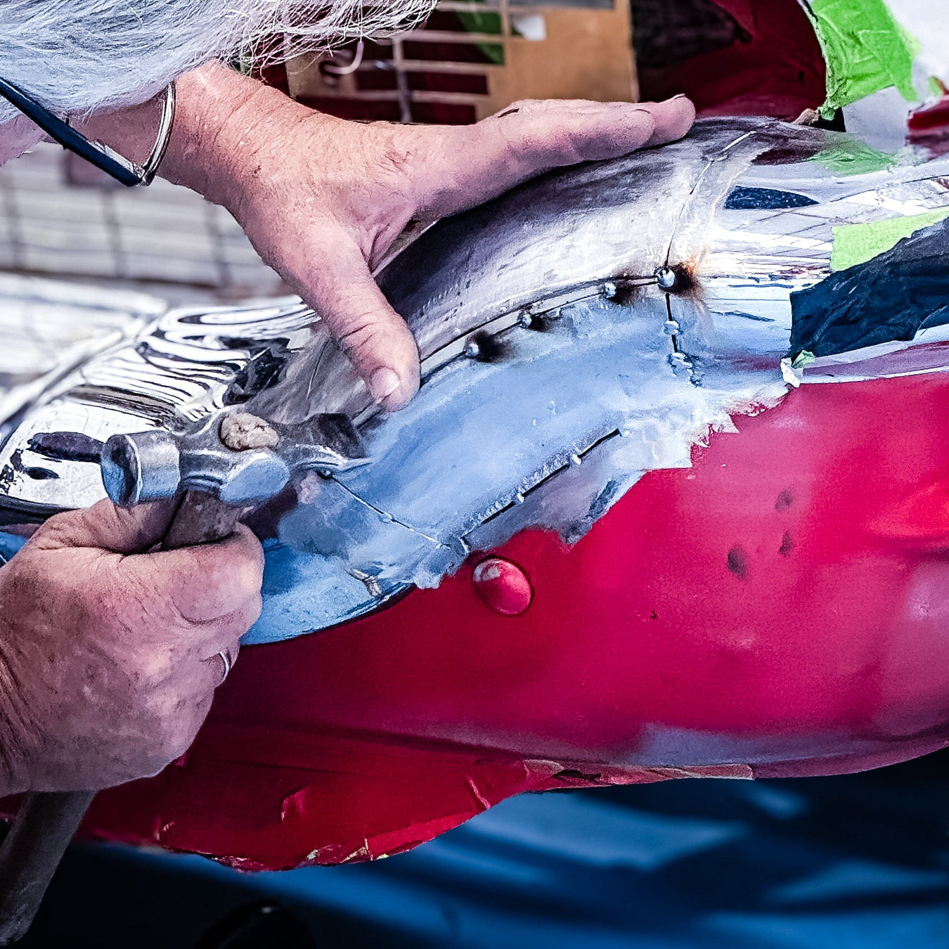 A mechanic with gray hair and glasses works on a red and white classic car body in a workshop setting.