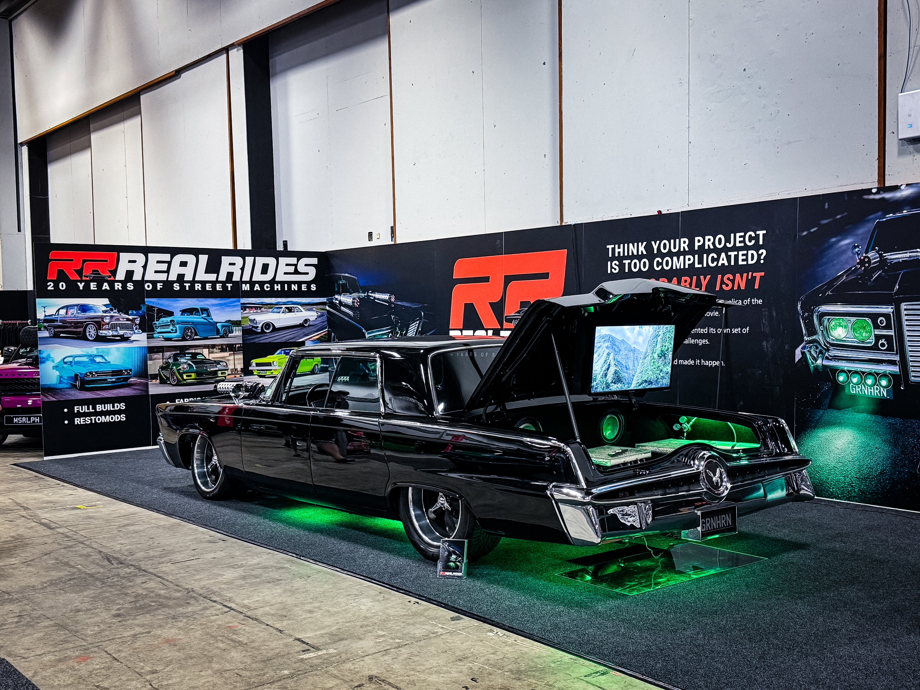 A customized black 64 IMPERIAL CHRYSLER BLACK BEAUTY REPLICA FROM THE GREEN HORNET MOVIE details and green underglow, parked on a carpeted display floor in front of a RealRides banner.