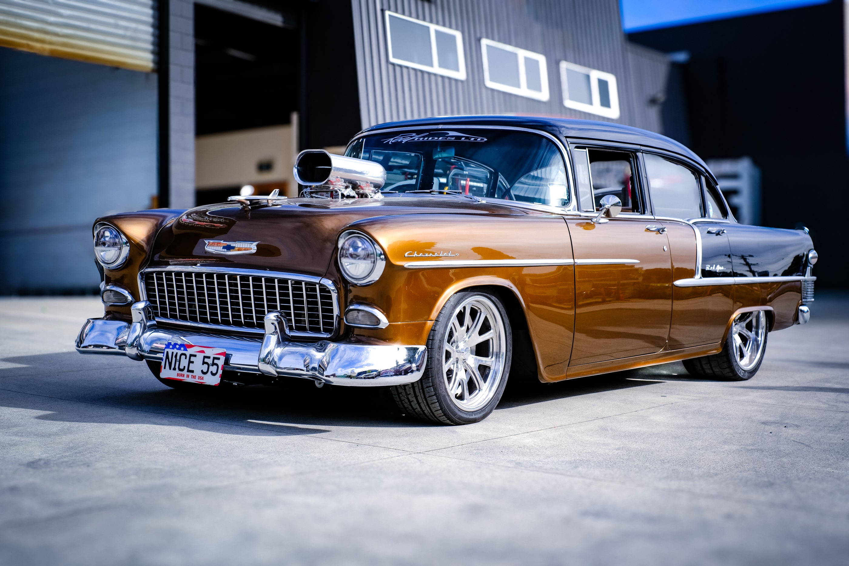 A customized 1957 Chevrolet 2100 in metallic bronze-brown with a black roof, chrome custom wheels, and an exposed supercharger, parked in front of a garage.