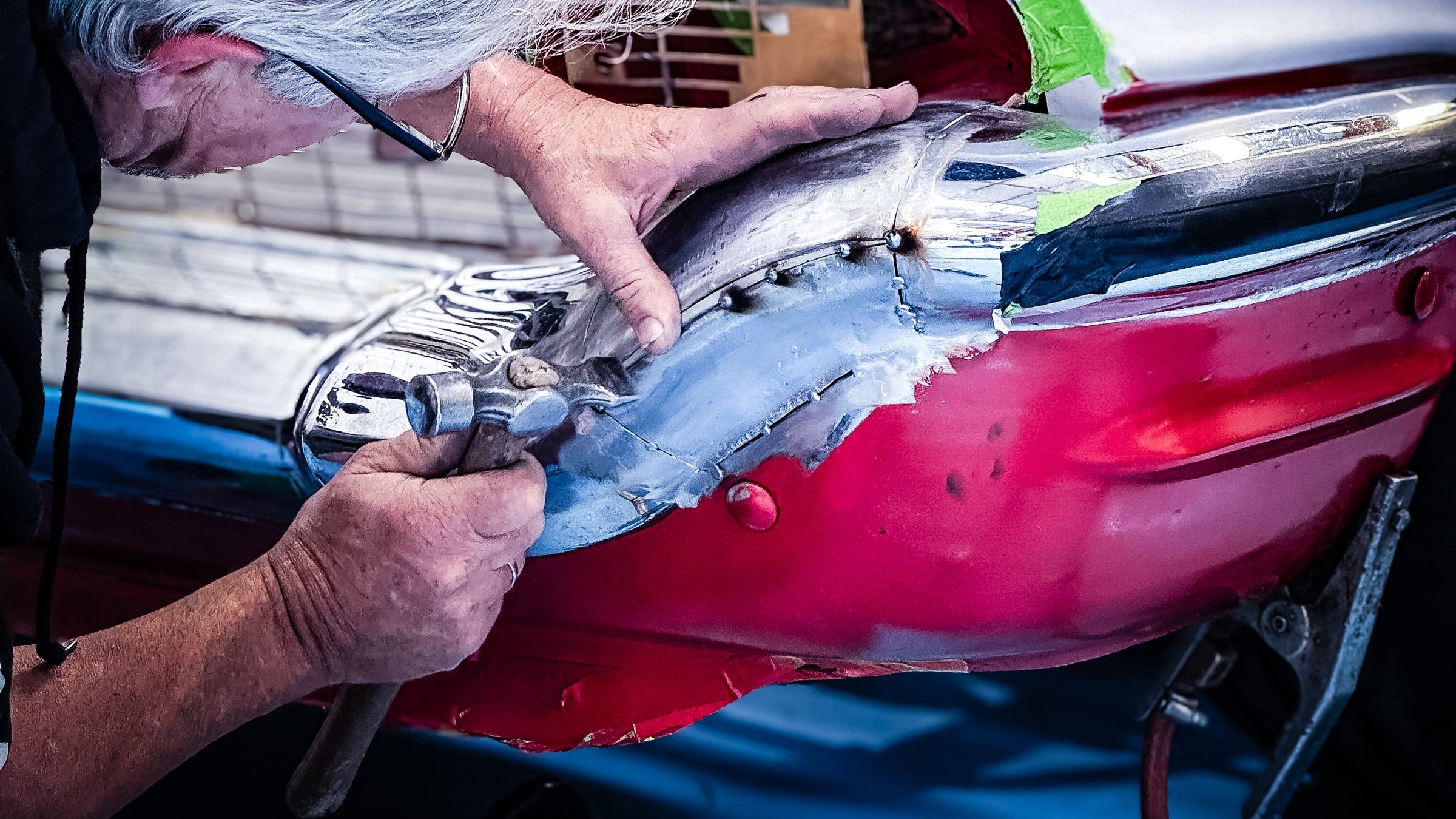 A mechanic with gray hair and glasses works on a red and white classic car body in a workshop setting.