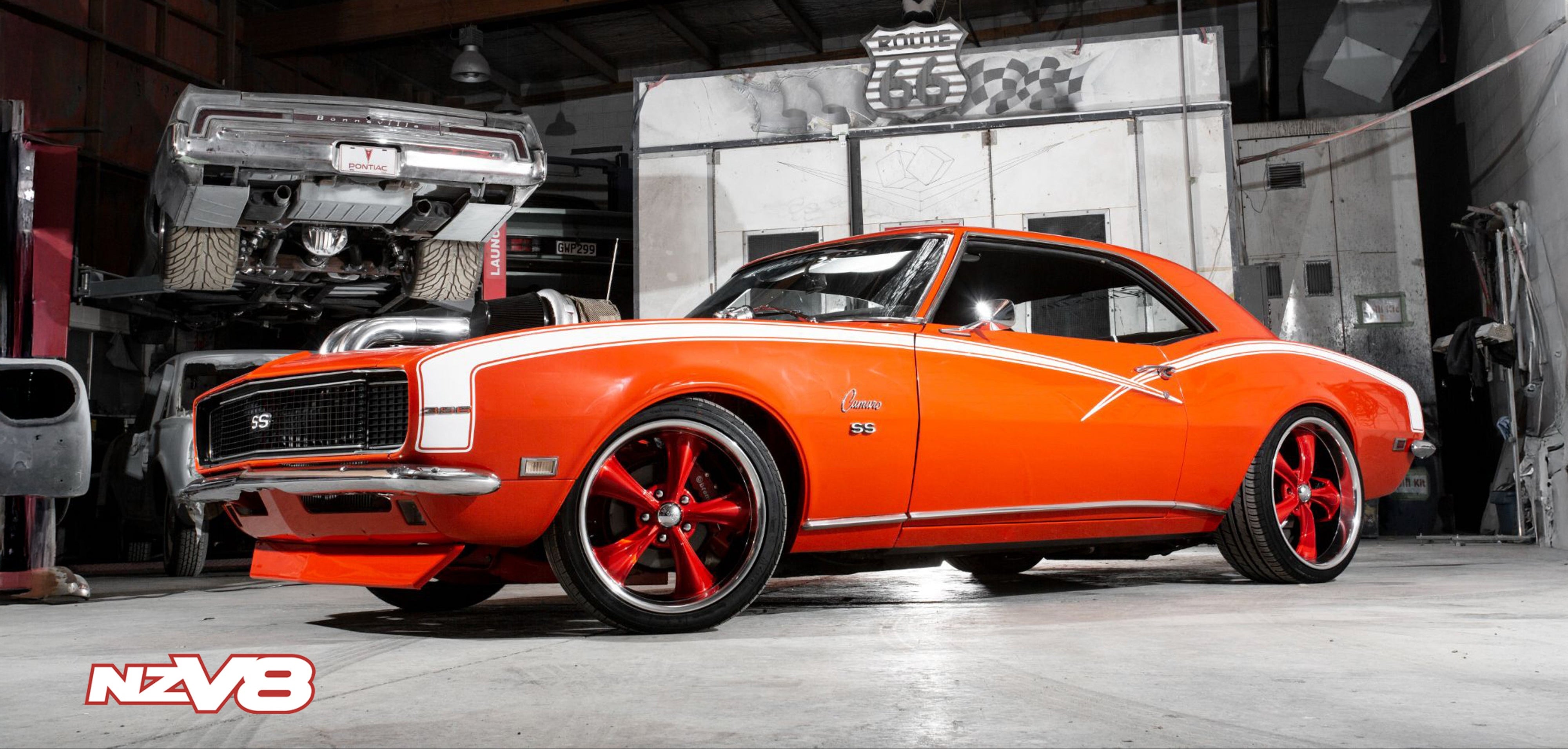 A bright orange classic muscle car with red multi-spoke wheels parked inside a garage workshop. The car features a white racing stripe along the side and a chrome grille.