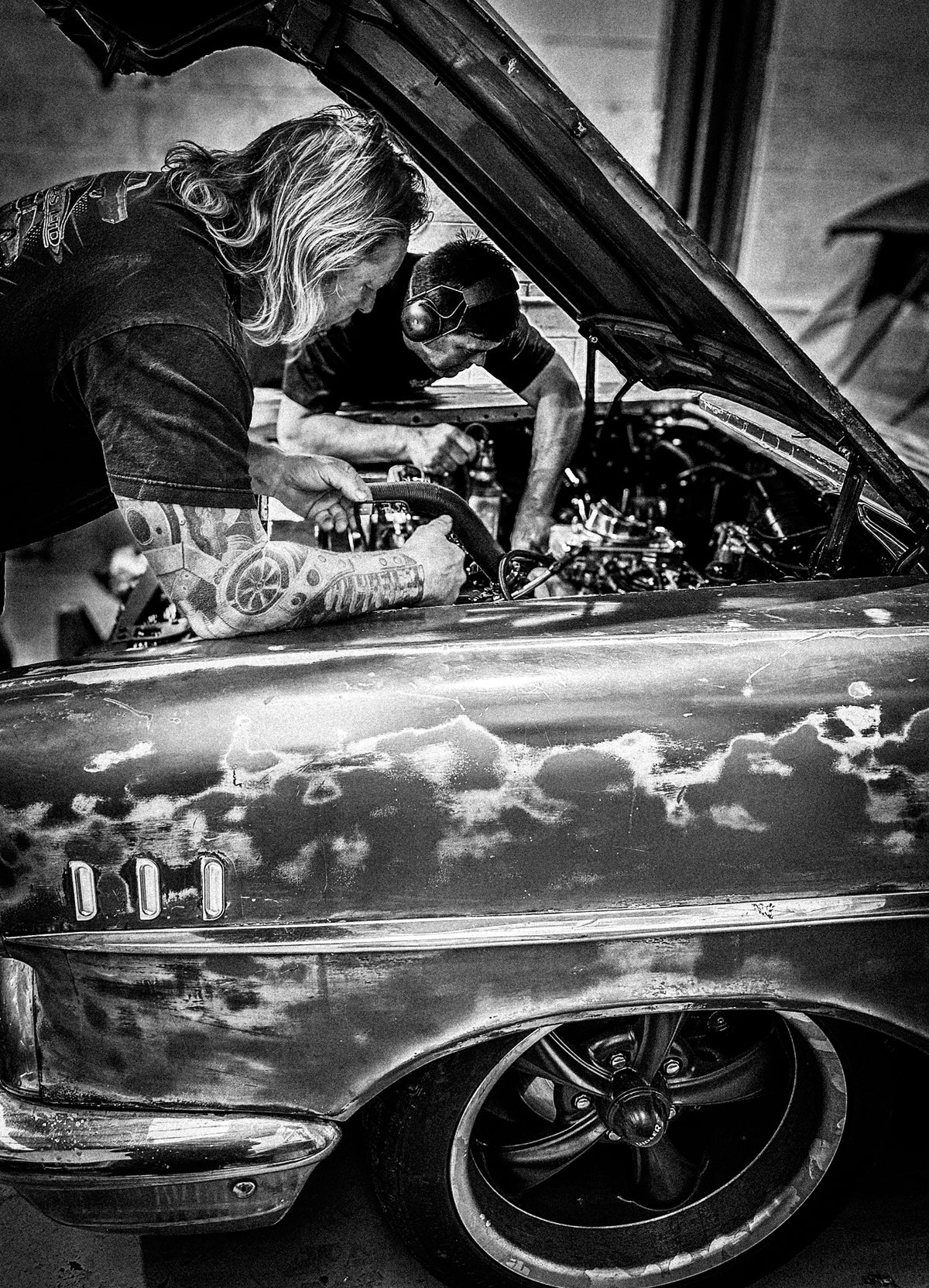 Black and white photo of two men working under an open car hood in a garage, with a custom, paint-splattered classic car in the foreground.