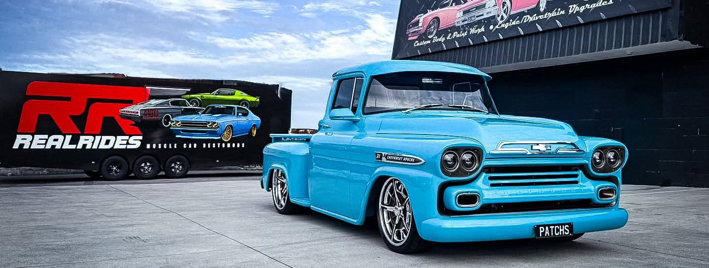 A bright turquoise classic Chevrolet pickup truck is parked on a concrete lot in front of a large RealAides trailer and branded wall sign. The truck is shown in a real outdoor setting with sky, pavement, and vehicle trailers visible, so this is Lifestyle rather than Product/Packaging.