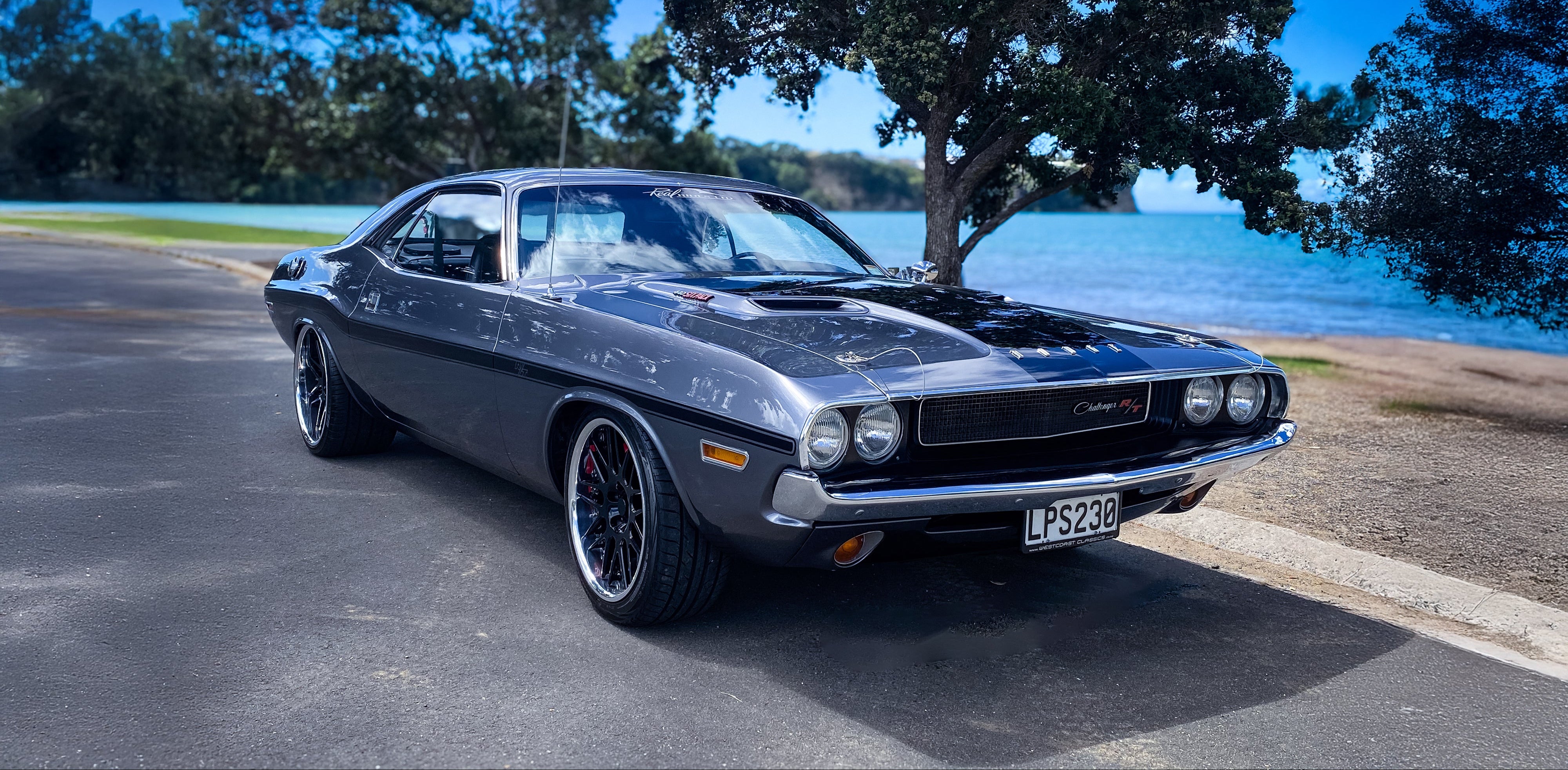 A dark metallic gray classic muscle car parked on an asphalt road next to a lake with trees and blue sky in the background.